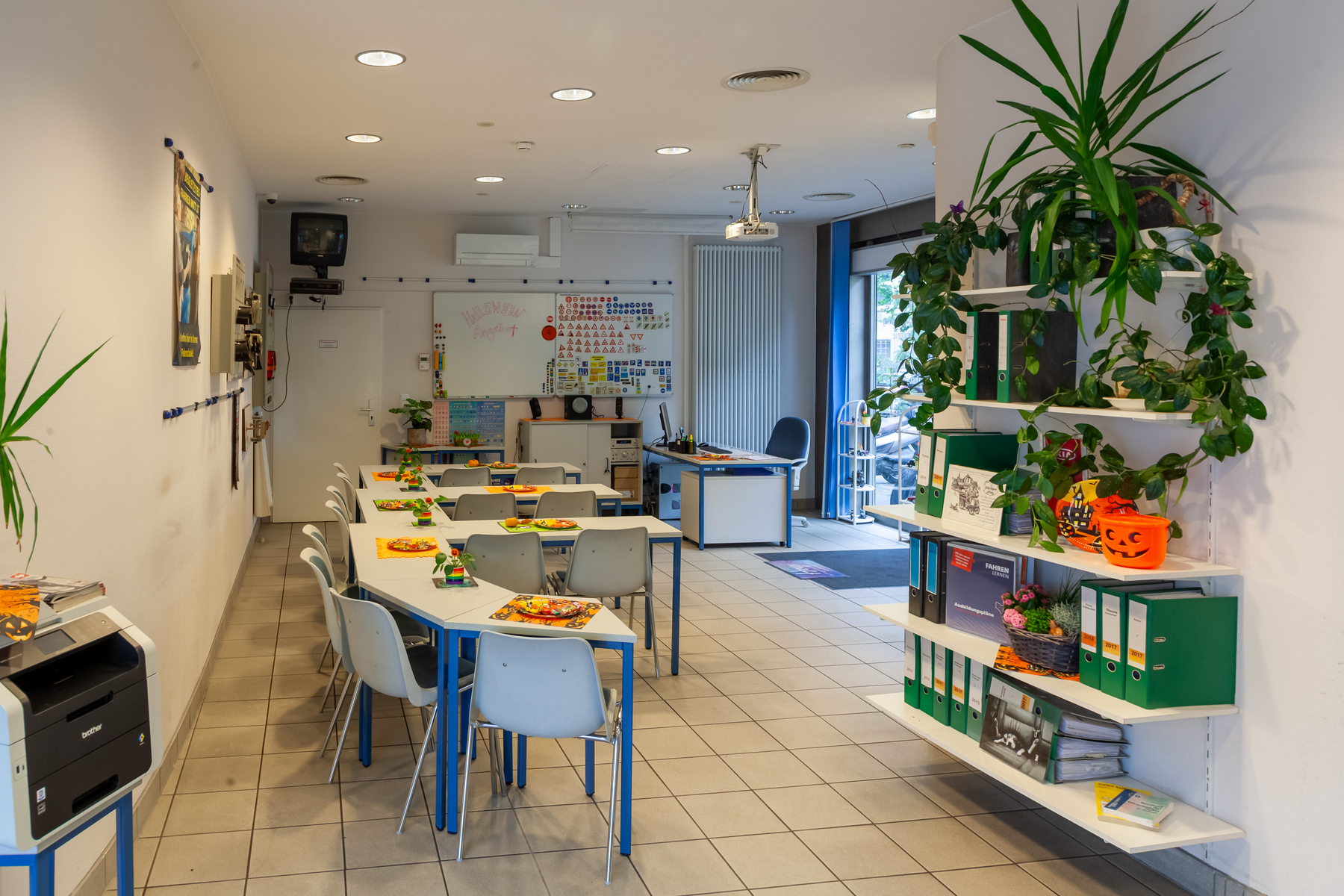 Ein Klassenzimmer mit mehreren U-förmig angeordneten Tischen, die jeweils mit Tellern und Tassen ausgestattet sind. Um die Tische herum stehen Stühle, eine Tafel, bunte Dekorationen, Pflanzen in Regalen und ein Schreibtisch im hinteren Teil des Raumes, der durch eine Glastür zu erreichen ist.