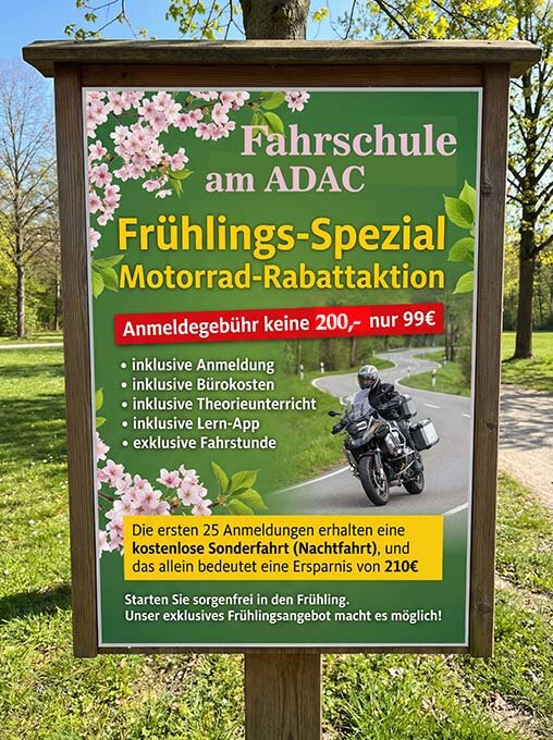 ein schild am straßenrand unter bäumen wirbt für eine frühjahrsaktion einer fahrschule: "motorrad rabattaktion". auf dem schild sind blumen, sonderpreise, eine liste der inbegriffenen dienstleistungen und ein foto einer person auf einem motorrad abgebildet.